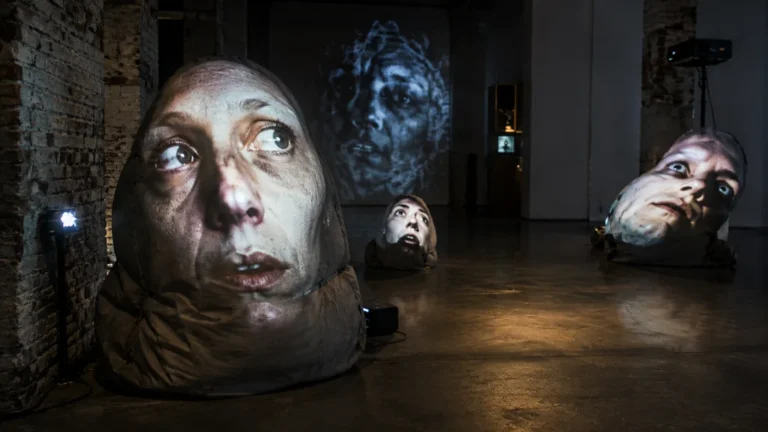 Installation d’art vidéo contemporain avec des visages projetés sur des sculptures dans une galerie sombre